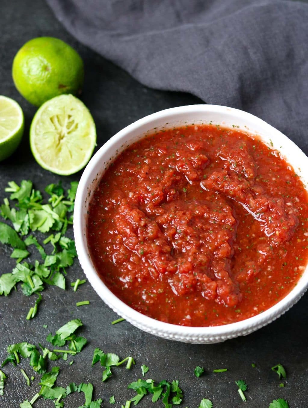 Restaurant Style Salsa in a white bowl with cilantro scattered on the counter and limes in the background