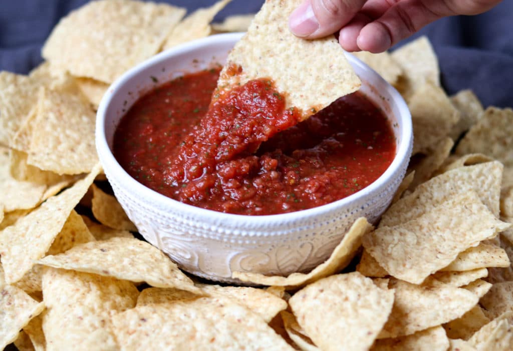 Restaurant Style Salsa in a white bowl surrounded by chips, a tortilla chip is being dipped into the salsa