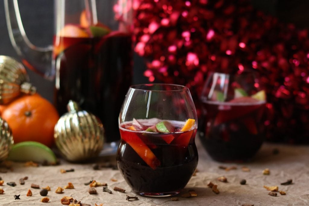 A glass of Red Wine Winter Sangria mixed with oranges and apples. Holiday decor and a pitcher of winter sangria are in the background. 