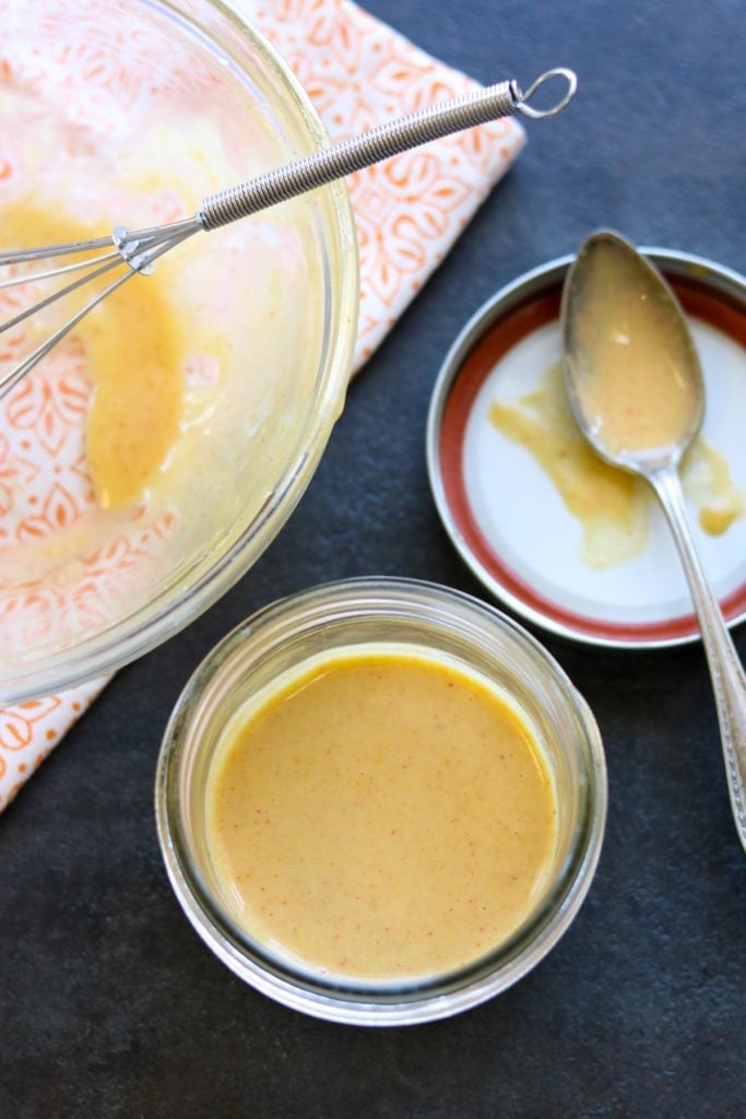 A mason jar of honey mustard dipping sauce, a mixing bowl with a whisk is sitting on a napkin to the side