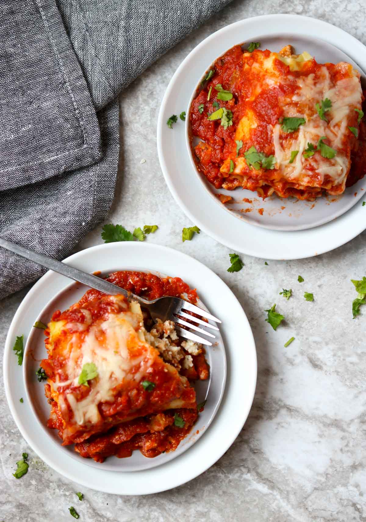Two lasagna roll ups on white plates with one fork and a gray napkin