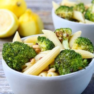 penne in a bowl with broccoli and pine nuts, lemons in the background
