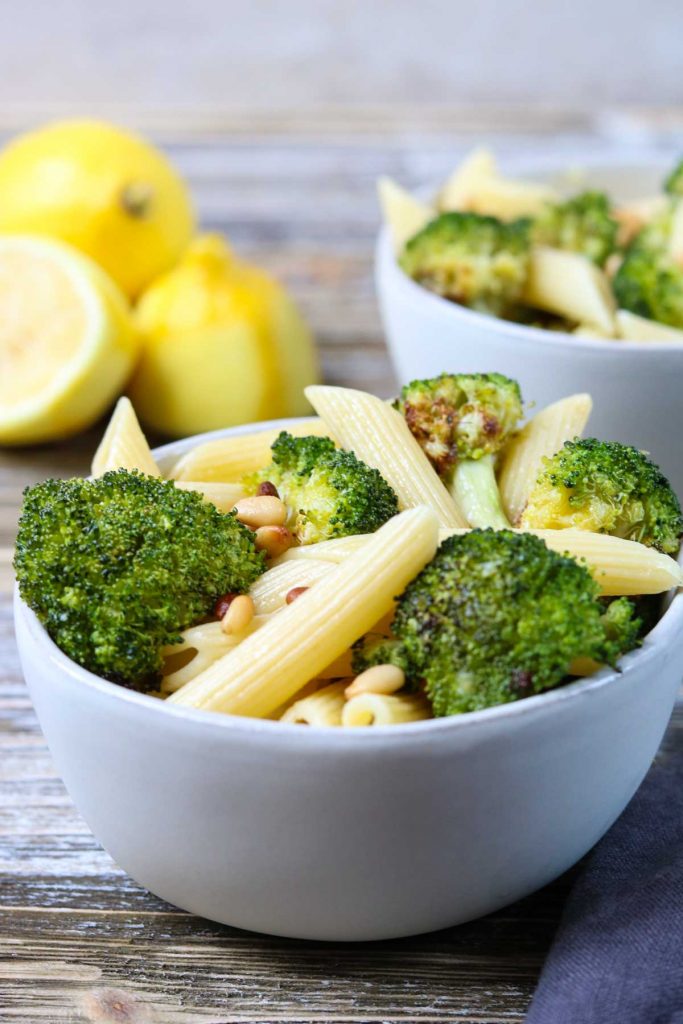 A white bowl full of penne pasta, broccoli, lemon, and pine nuts