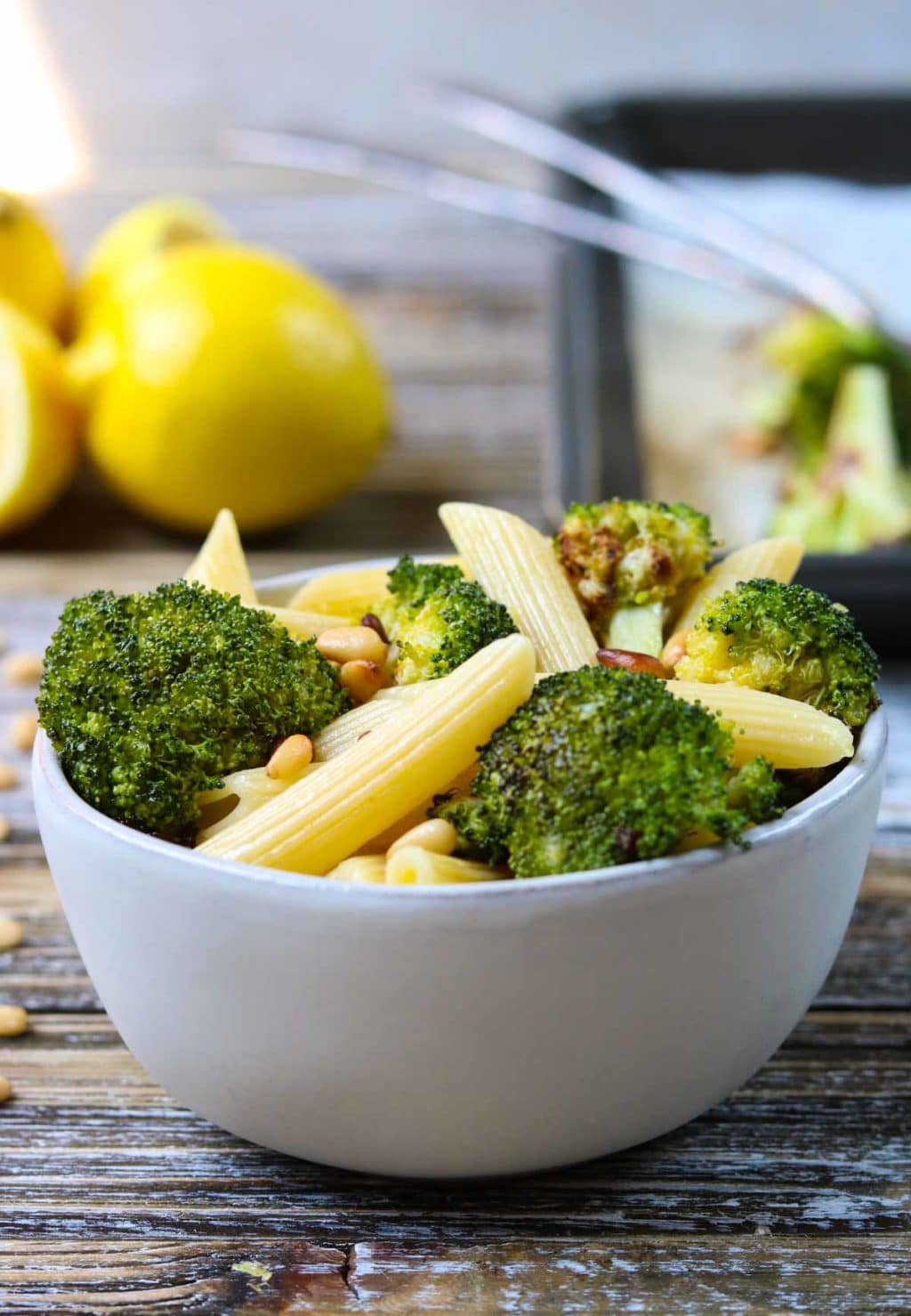 A white bowl full of pasta and broccoli, lemons in the background