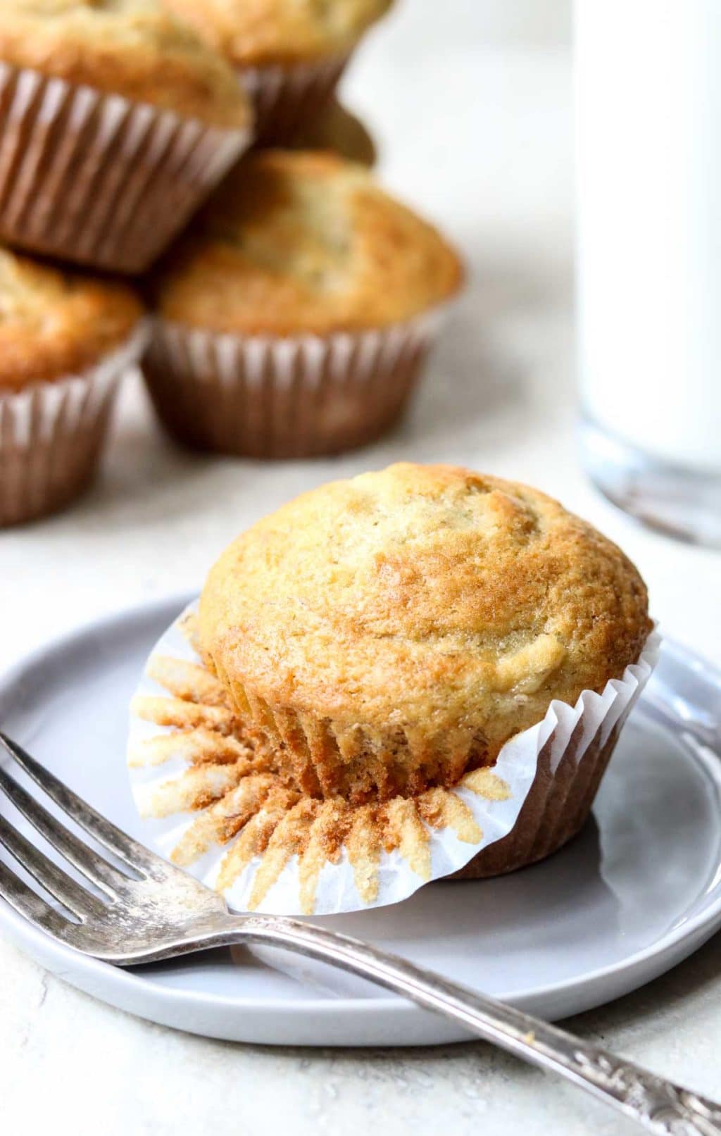 A banana bread muffin with the paper liner peeled down half way, sitting on a gray plate with a fork