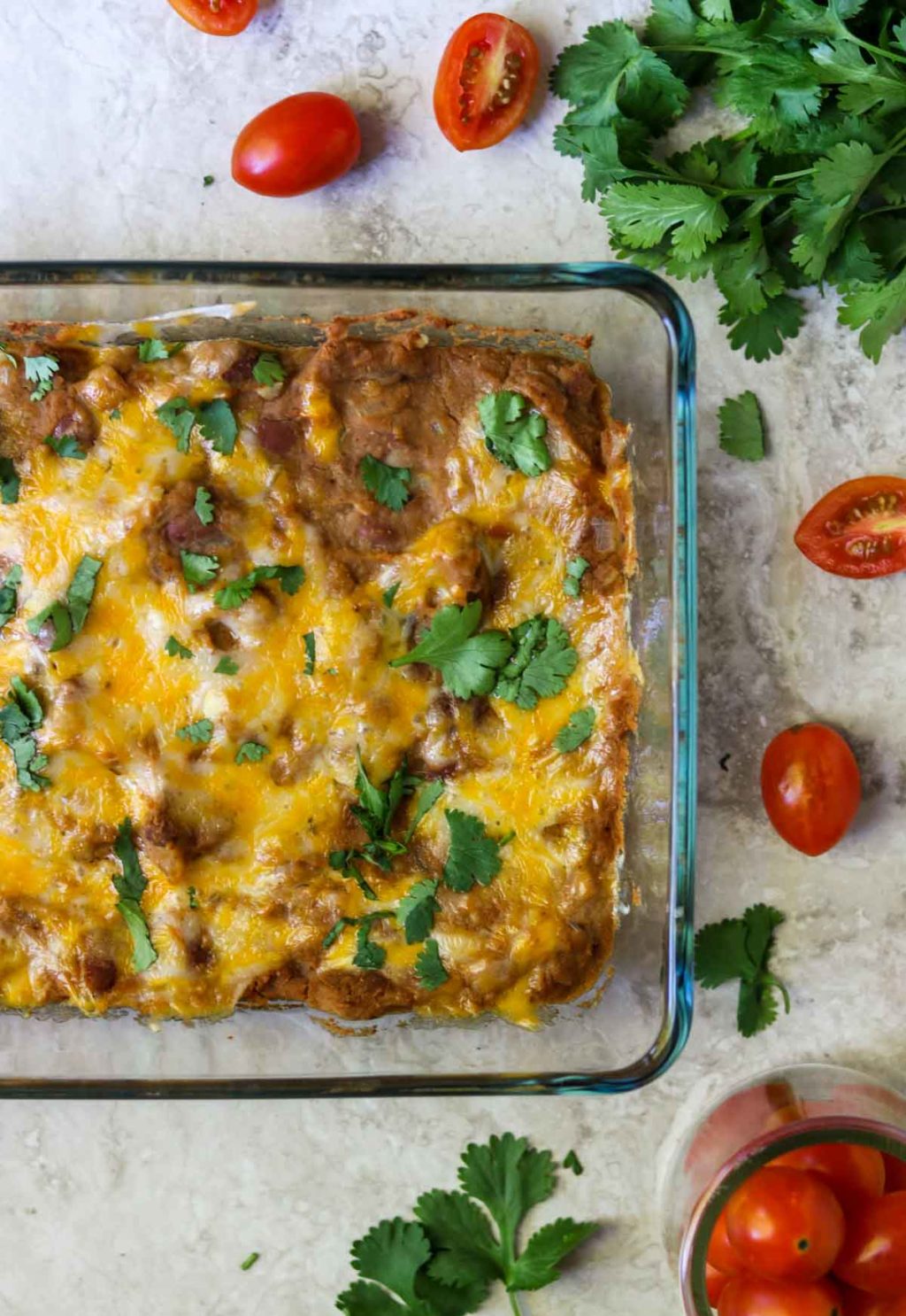 A baking dish of refried beans topped with cheese and cilantro. tomatoes and cilantro scattered on the counter around the baking dish