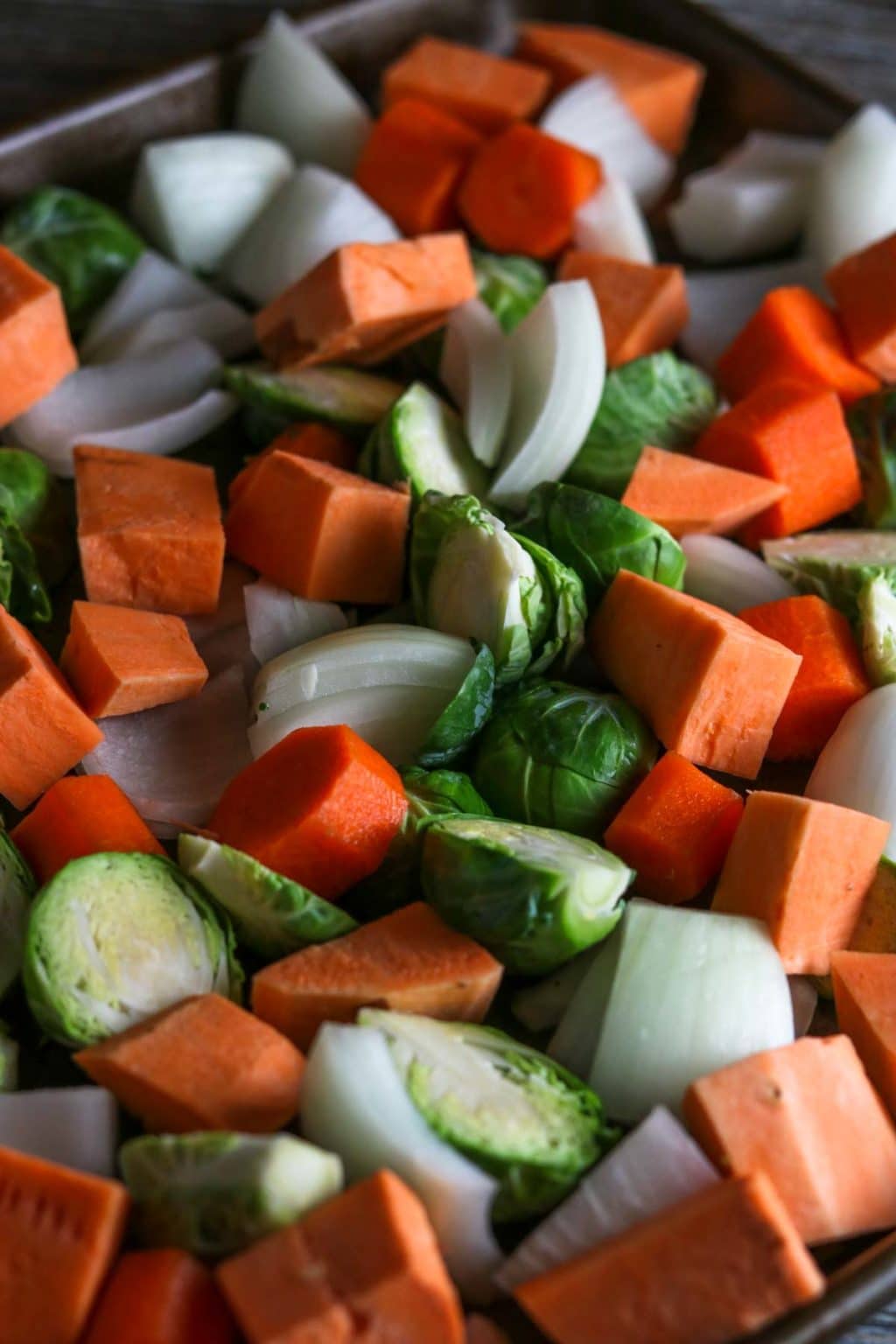 A sheet pan full of raw, cut veggies: brussel sprouts, onions, sweet potatoes, and carrots