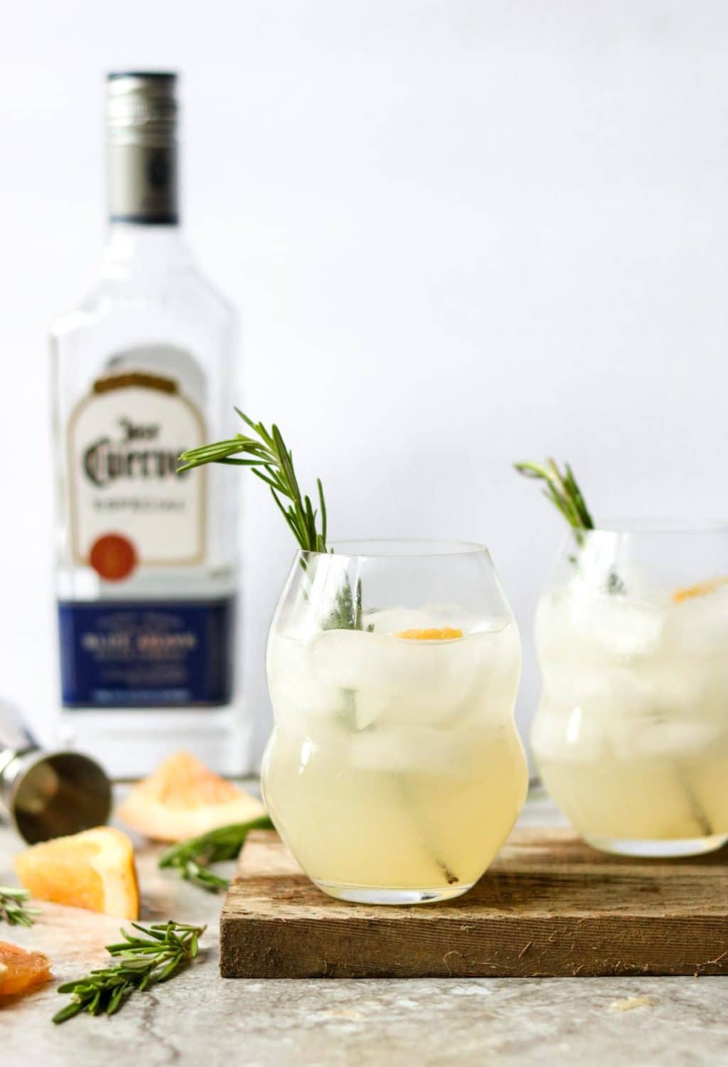 two glasses of Rosemary Paloma with rosemary sprig garnish, a bottle of tequila in the background