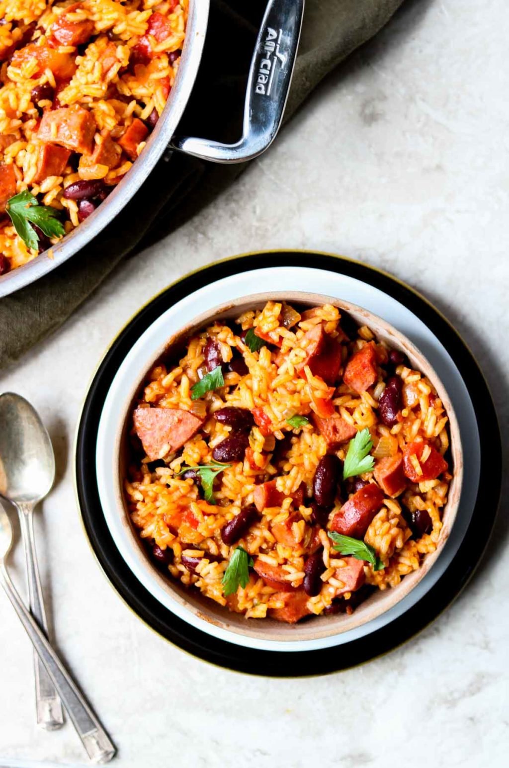 A bowl of dirty rice and beans with two spoons to the side, an all clad skillet at the top of the picture