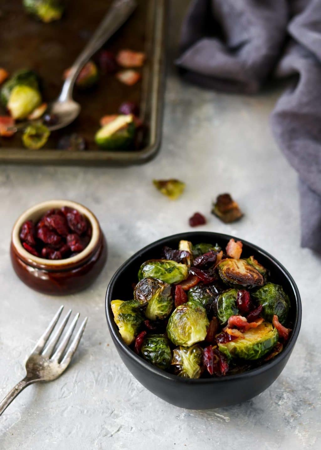 brussels sprouts in a black bowl with a cookie sheet in the background and a small bowl of dried cranberries