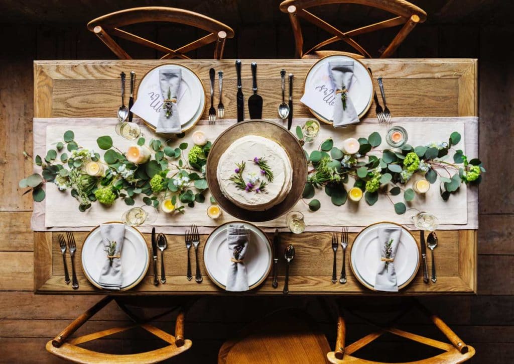 wooden dinner table set with white plates and blue napkins, with a white table runner and greenery and candles