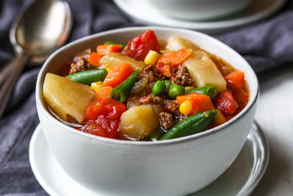 a white bowl of easy instant pot hamburger and vegetable soup- potatoes, tomatoes, beef, peas, corn, carrots, and green beans.