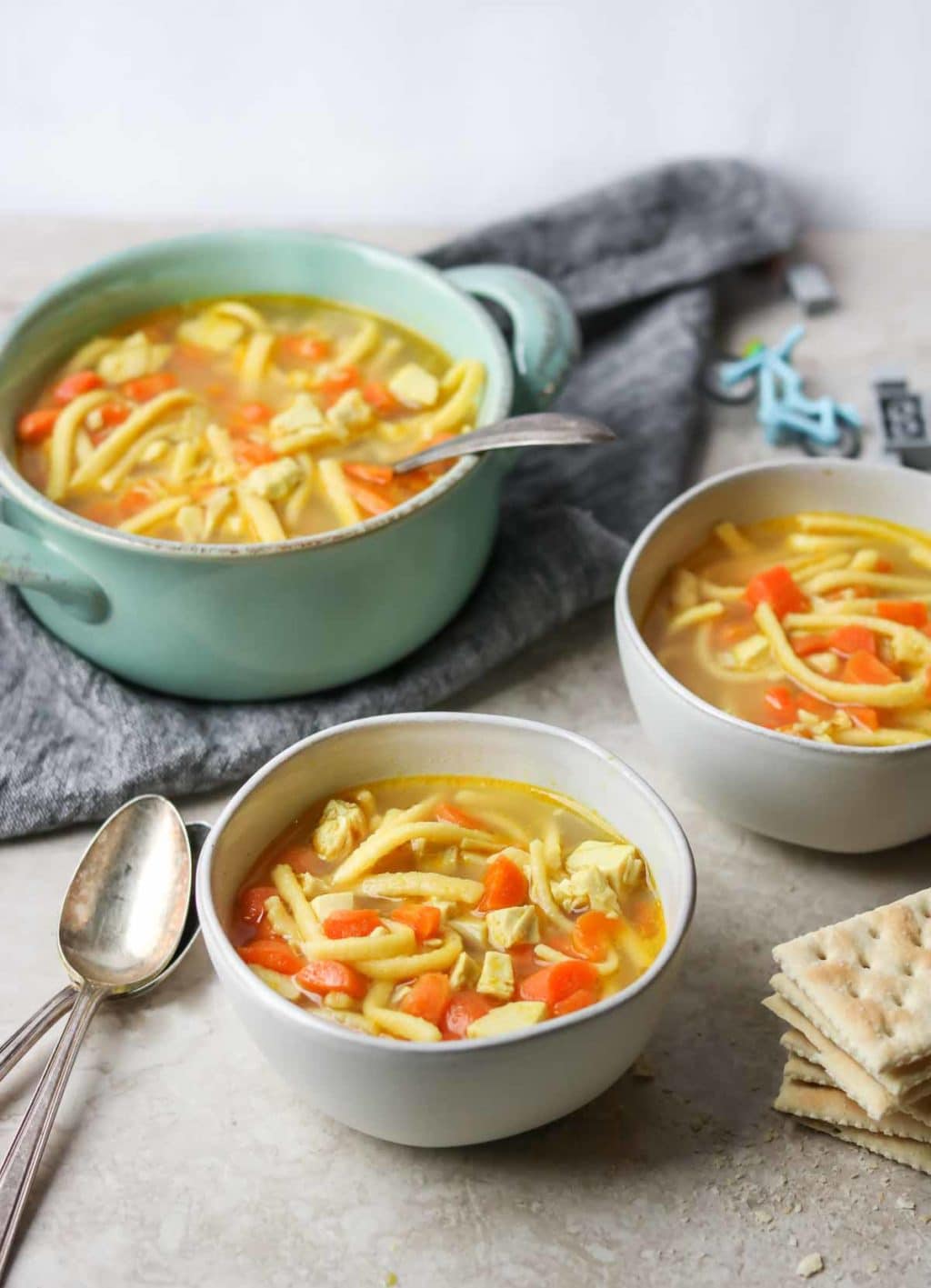 two white bowls of kid friendly chicken noodle soup with two spoons, a large pot of chicken noodle soup sitting on a towel