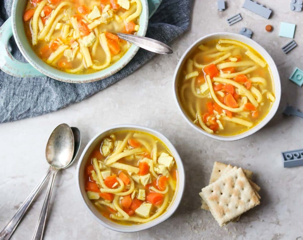 Two bowls of chicken noodle soup for kids, a pile of crackers, and a few legos scattered around