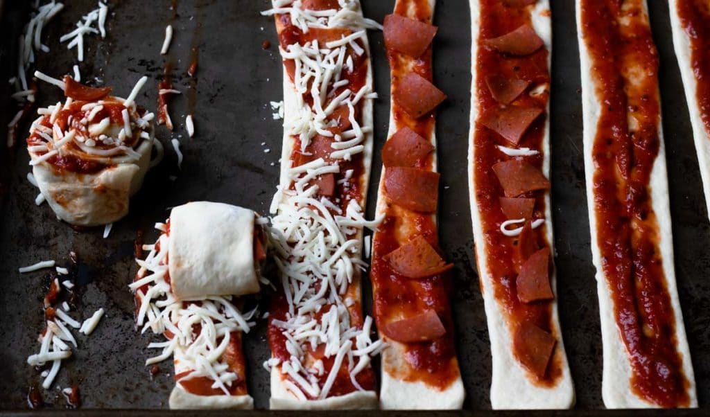 pizza roll ups on a baking sheet topped with marinara, pepperoni, and cheese, rolled up