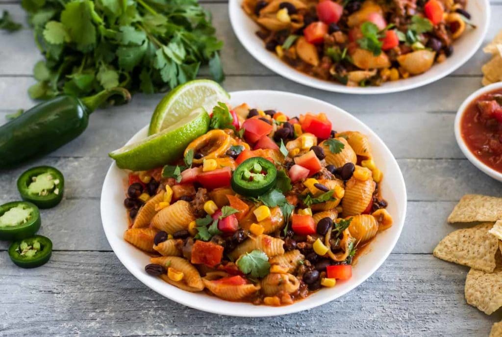 a white bowl of pasta with ground beef, corn black beans, peppers tomatoes, and garnished with limes, cilantro and jalapenos