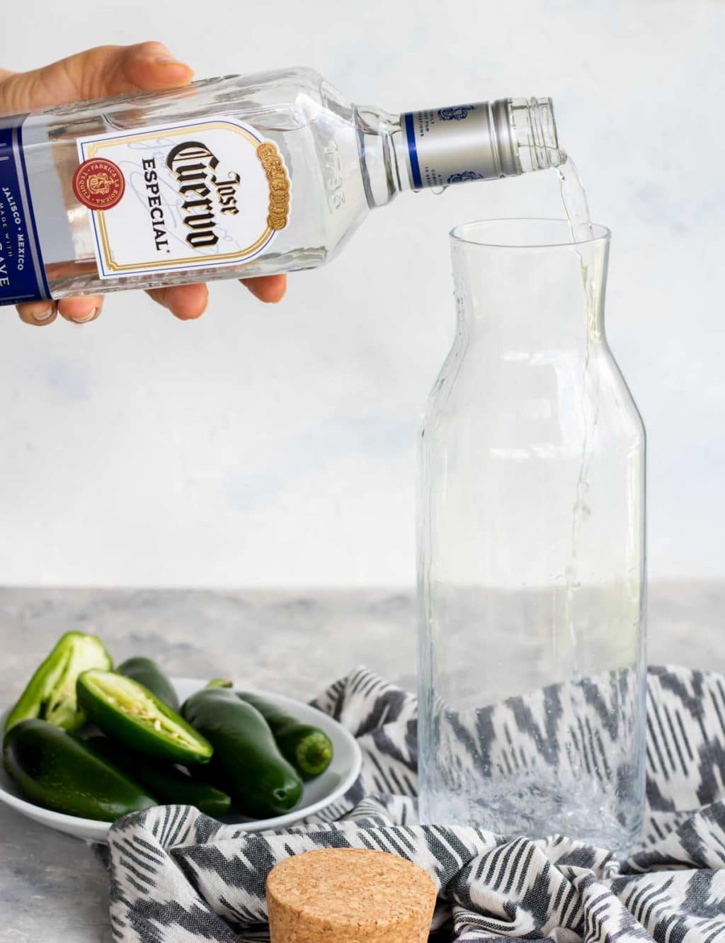 Pouring a bottle of silver tequila into a glass container with a bowl of jalapenos to the side