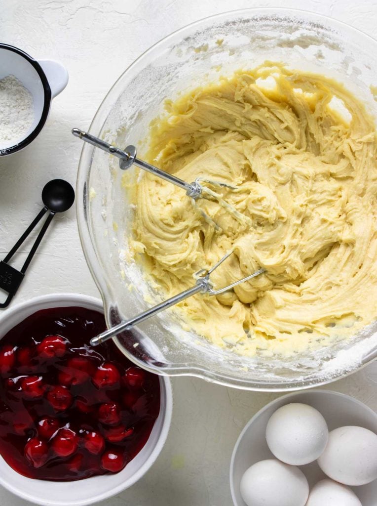 Cherry pie bar batter in a mixing bowl, cherry pie filling to the side with egg and flour