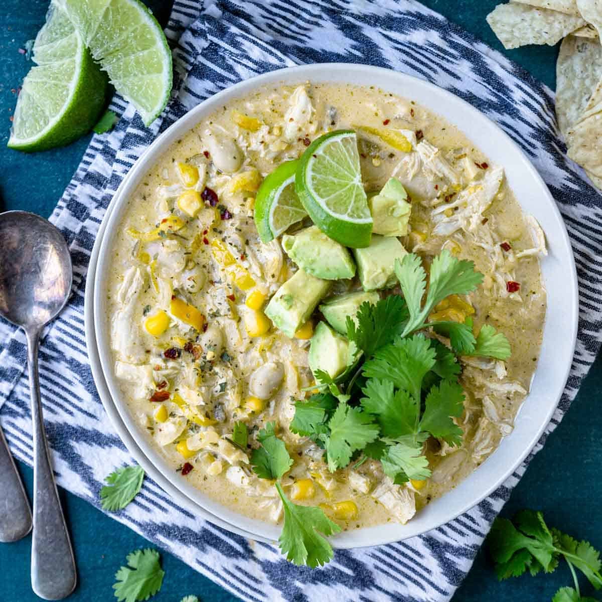 a bowl of white chicken chili topped with avocado, lime wedges and cilantro