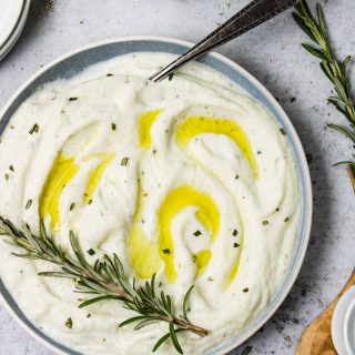 Cauliflower Puree in a bowl with olive oil over top and a rosemary sprig