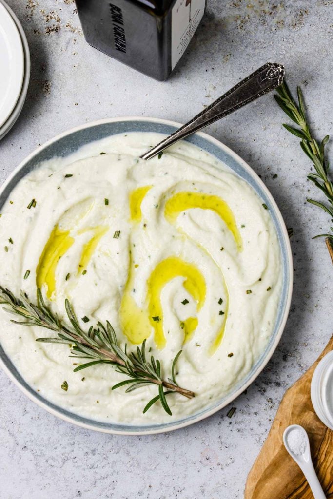 Cauliflower Puree in a bowl with olive oil over top and a rosemary sprig