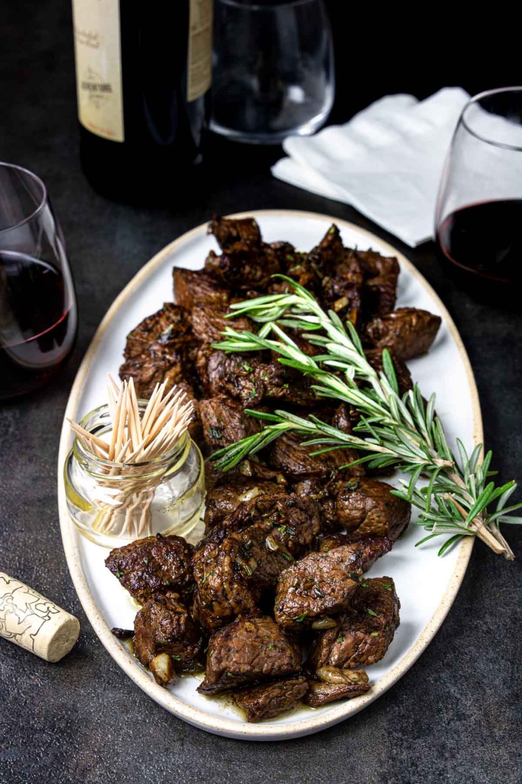Steak Bites with a garlic butter on a white plate, garnished with rosemary. A cup of toothpicks for serving as an appetizer.