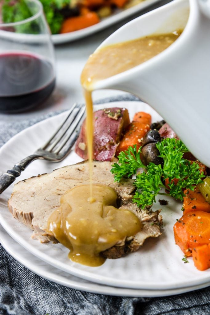 A slice of pork on a plate with vegetables and gravy being poured over top