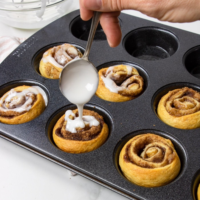 cinnamon rolls in a muffin tin being drizzled with frosting