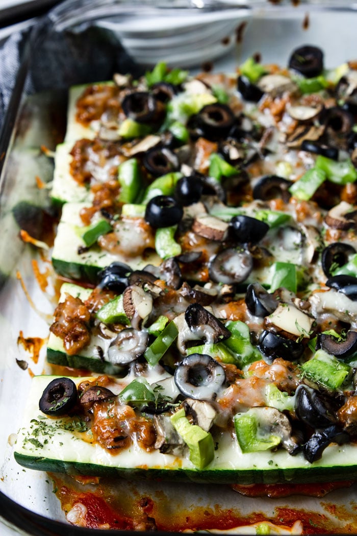 zucchini pizza boats in a glass casserole dish topped with black olives, green peppers, mushrooms and cheese