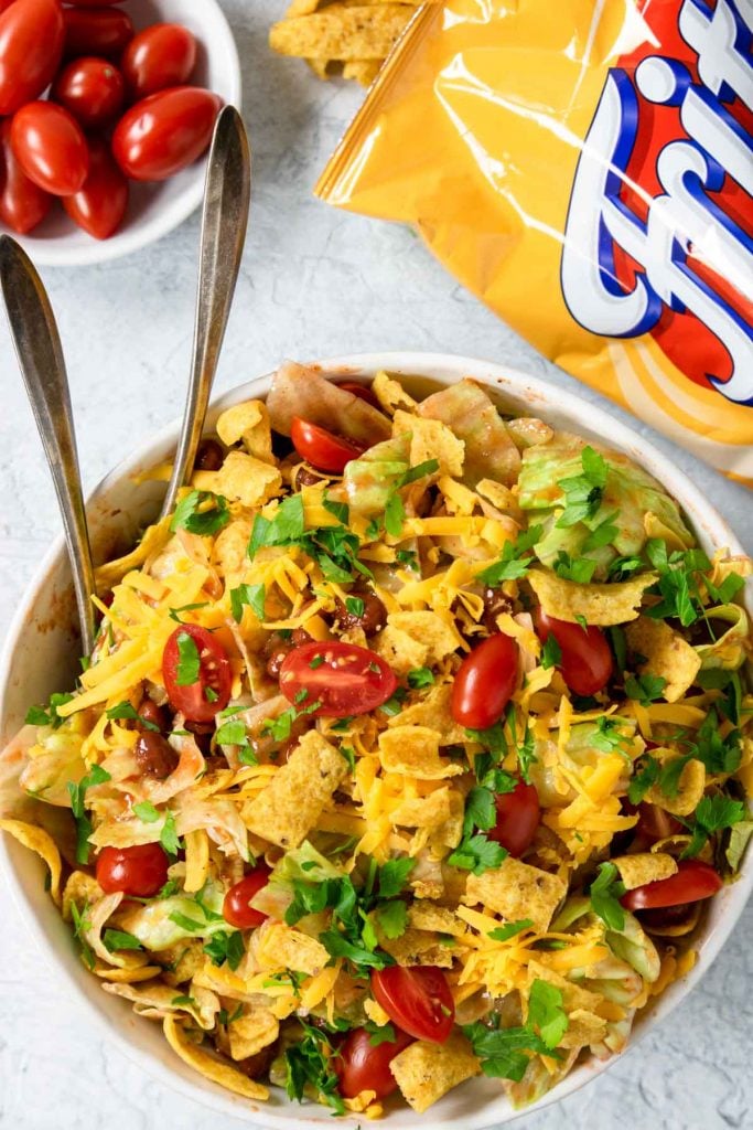 Large frito taco salad in a bowl with a bag of fritos and tomatoes to the side