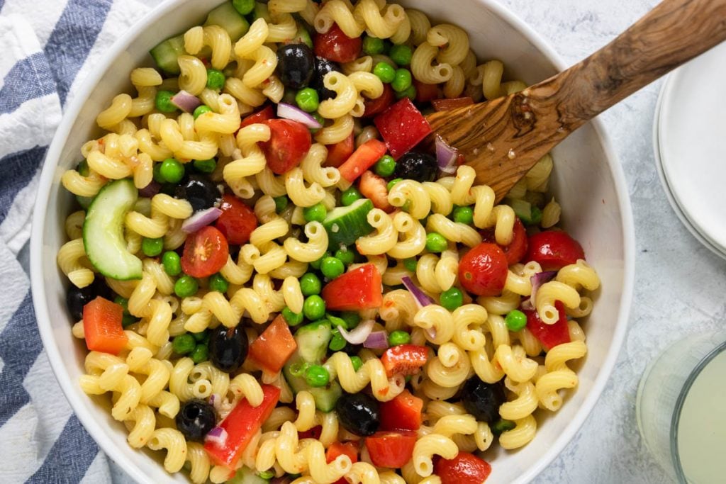 veggie pasta salad in a white bowl with cavatappi pasta and olives, peppers, tomatoes, cucumbers, and peas