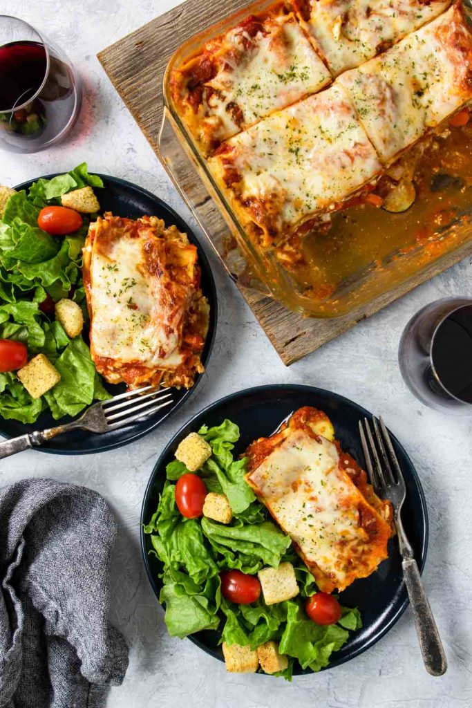 two blue plates with a slice of veggie lasagna and salad, and a glass baking dish of lasagna