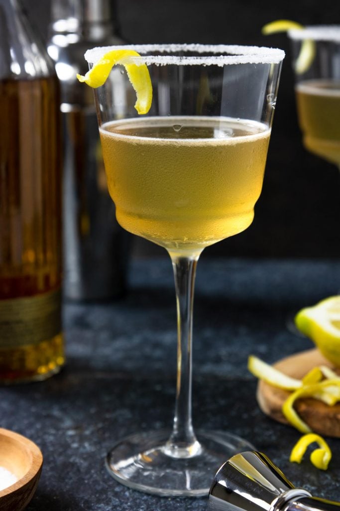 Anejo Sidecar in a stemmed glass with a sugar rim and lemon peel. Anejo tequila bottle to the side