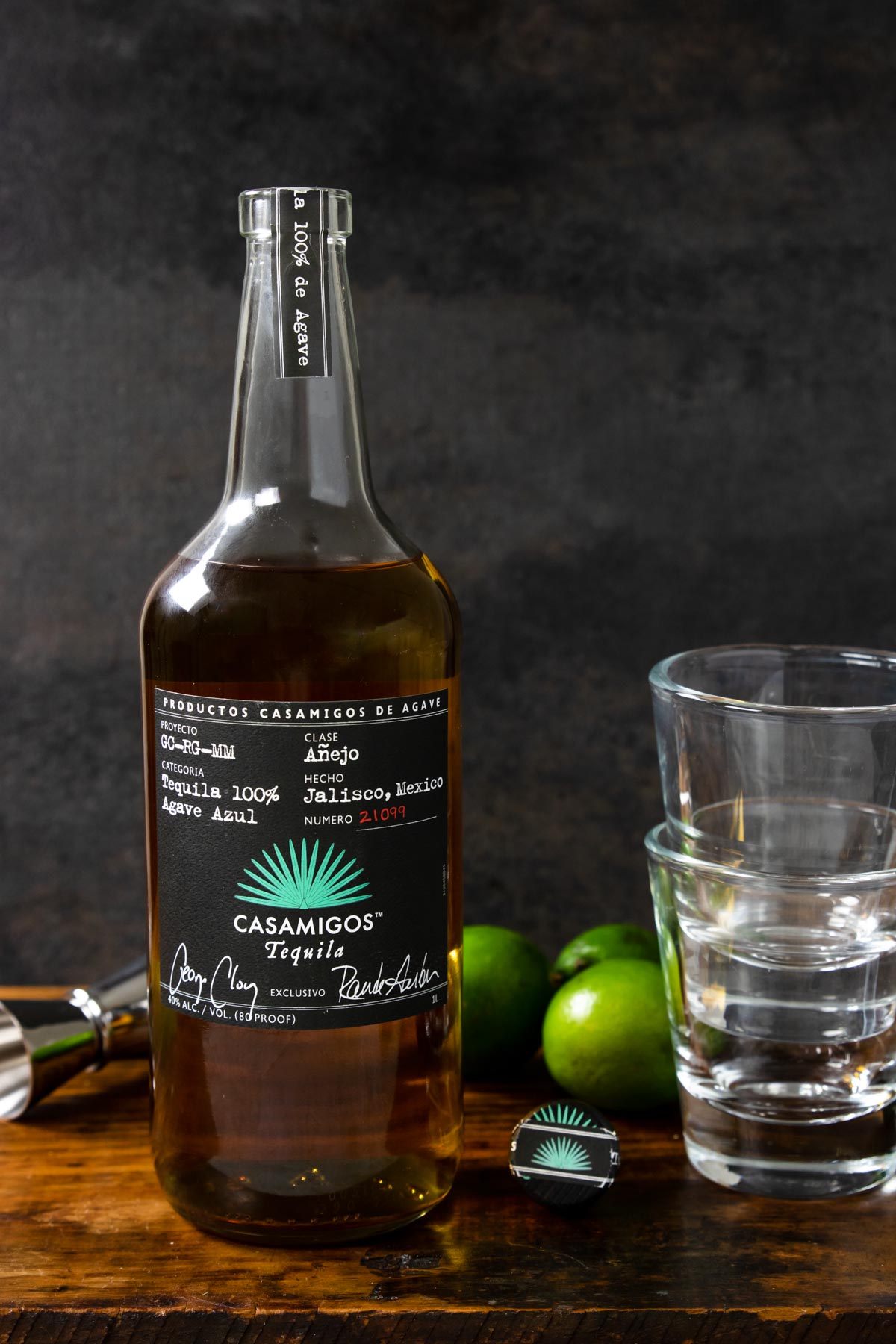 a bottle of casamigos anejo tequila, two glasses stacked, limes and a shot jigger