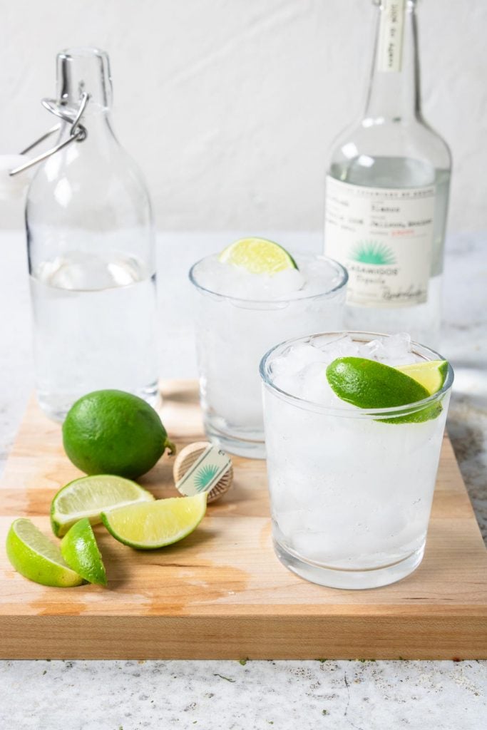 a cutting board with two cocktails, limes and a bottole of seltzer water. Also a bottle of Casamigos tequila in the background