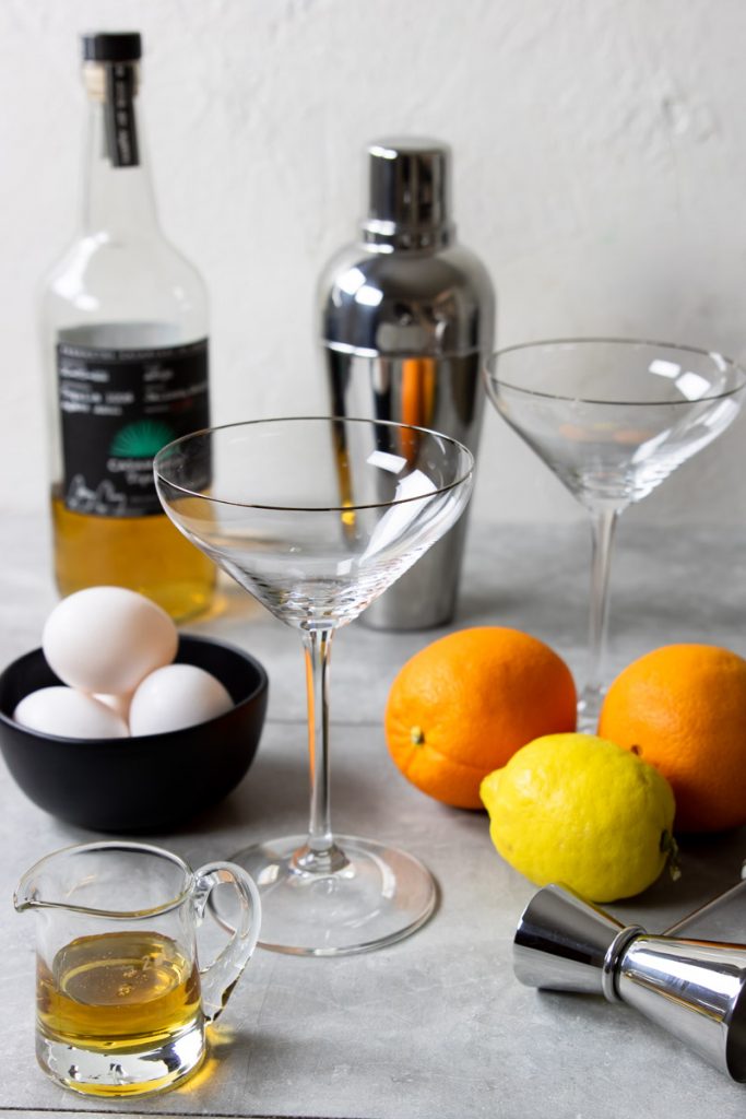 A line up of tequila sour ingredients - tequila, egg, lemon, oranges, agave syrup