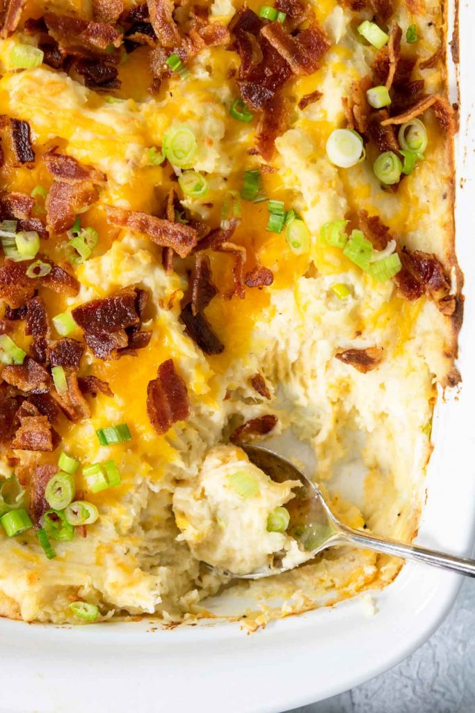 twice baked potato casserole in a baking dish topped with bacon and a spoon full taken out of the pan