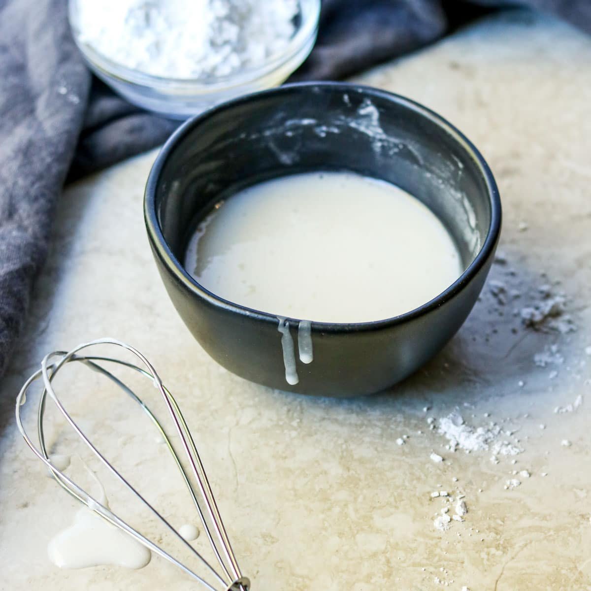 A black bowl of Powder Sugar Glaze with a whisk laying in front