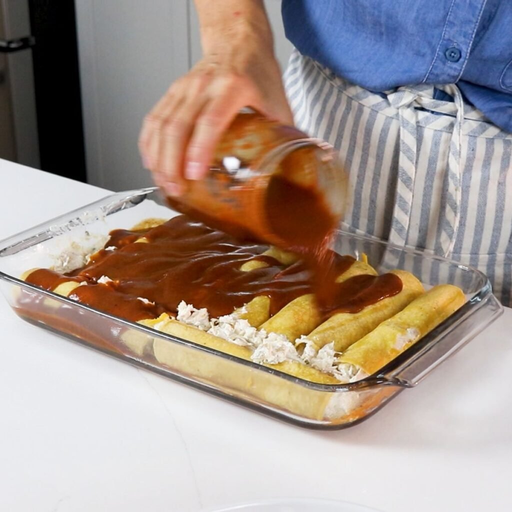 pouring red enchilada sauce over rolled chicken enchiladas with cream cheese
