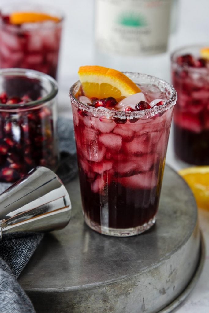 A glass of pomegranate margarita with a pomegranate and orange garnish