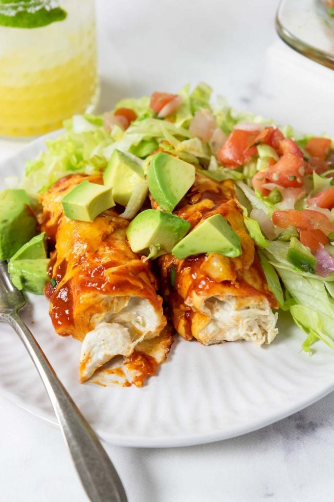 two enchiladas filled with creamy chicken on a plate with avocado, lettuce and pico de gallo