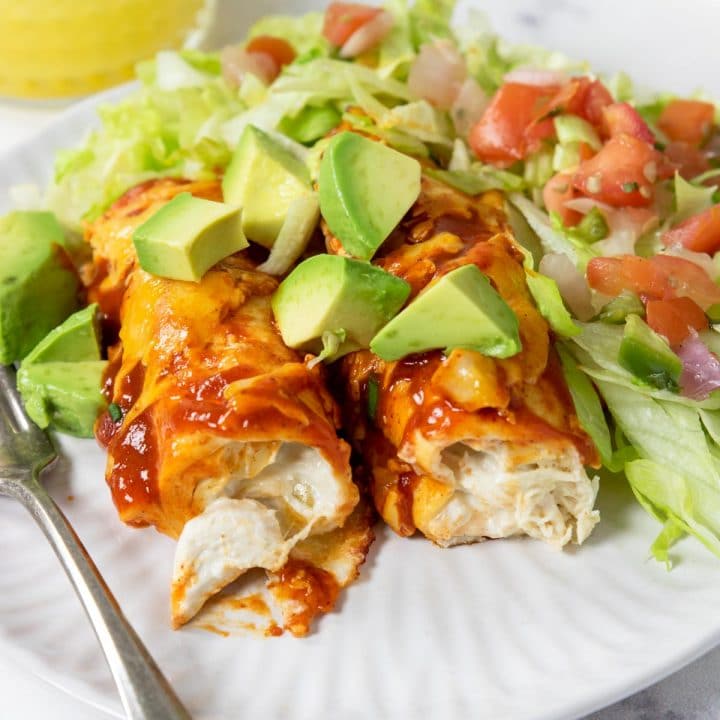 cream cheese chicken enchiladas on a plate with red chili sauce, cheese, and avocados
