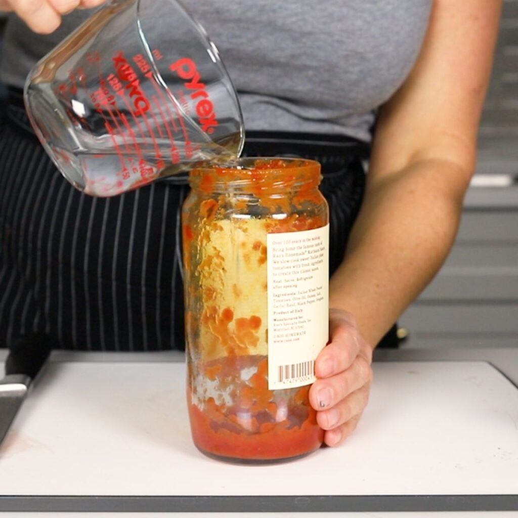 pouring water into a mostly empty jar of marinara 