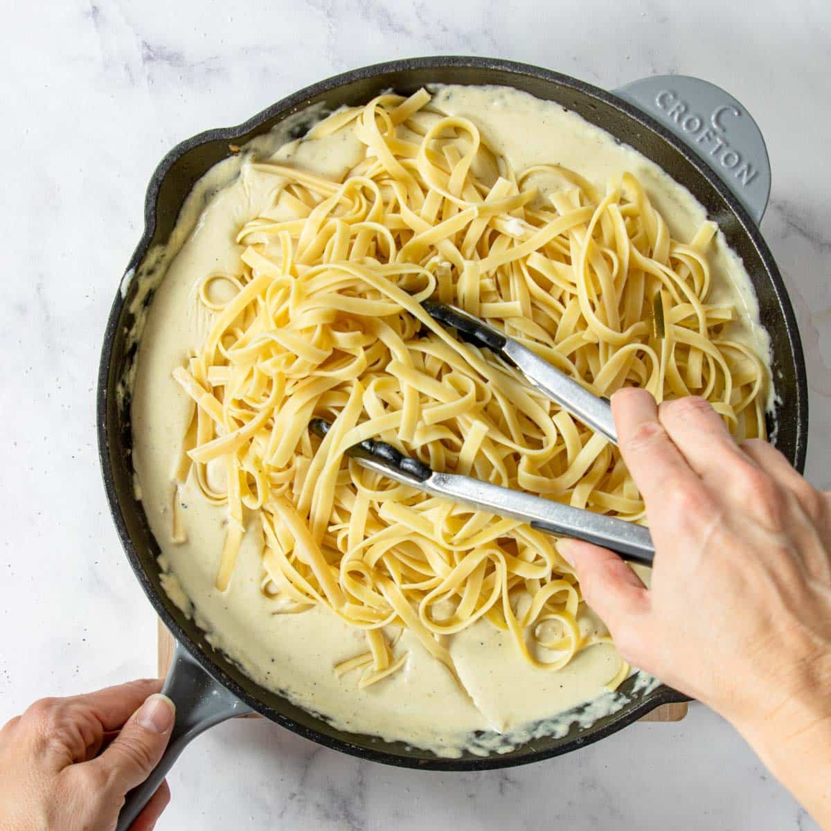 fettuccine pasta in a pan with Alfredo sauce, being tossed with tongs