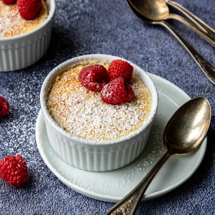 a white cup with lemon pudding cake topped with powdered sugar and raspberries
