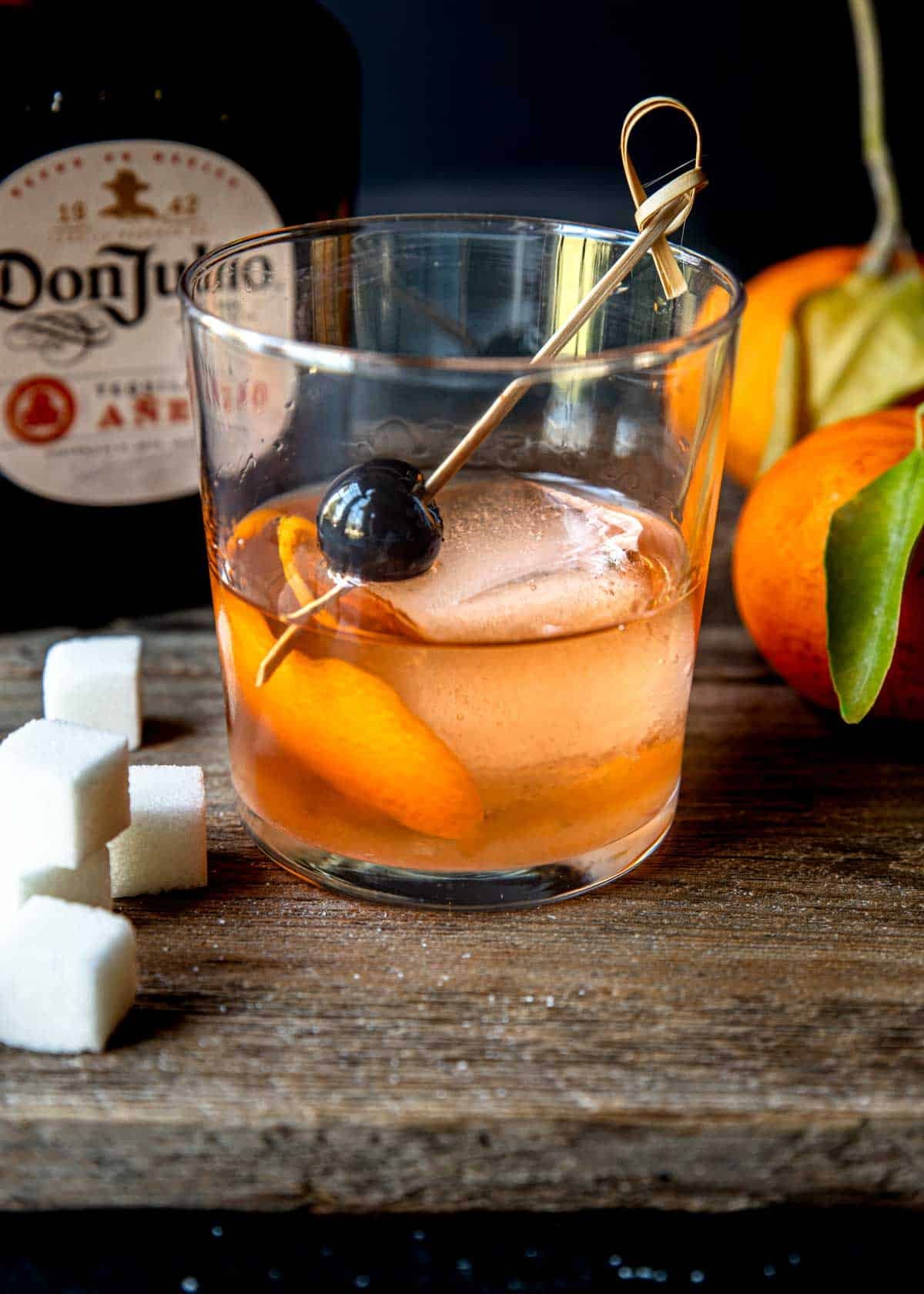 tequila old fashioned in a glass with a large ice cube, an orange peel and a cherry