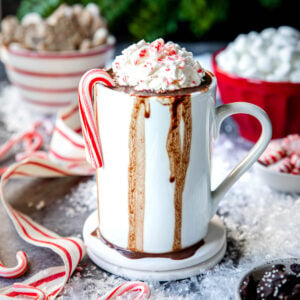 a mug of peppermint hot chocolate spilling over the edges with a candy cane on the side and whipped cream on top