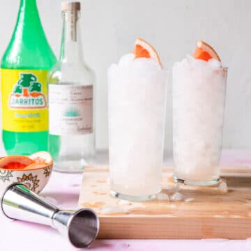 two grapefruit palomas with a bottle of grapefruit soda and silver tequila in the back