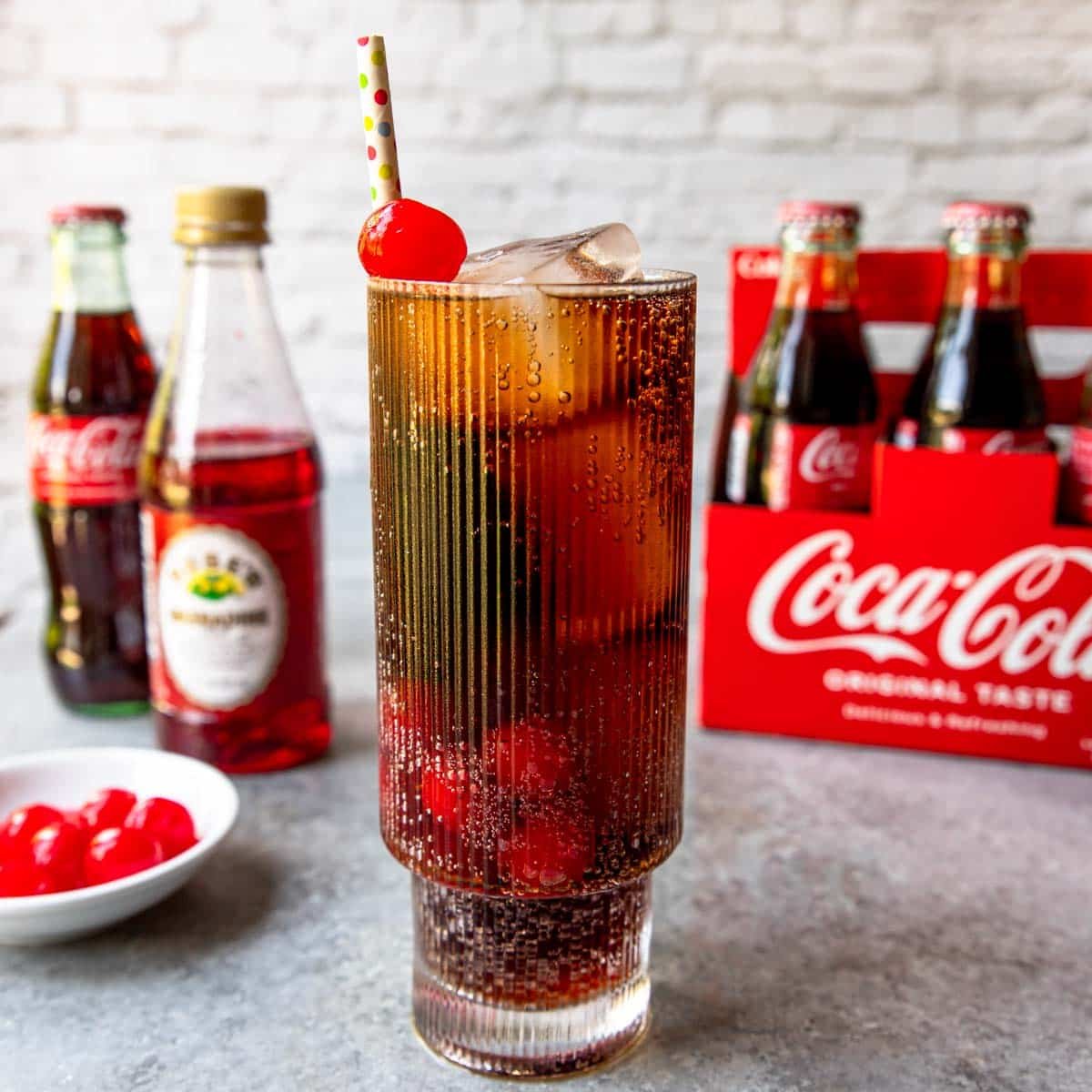 Roy Rogers cocktail (cherry coke) in a glass with a cherry on top and a straw, coca cola in the background