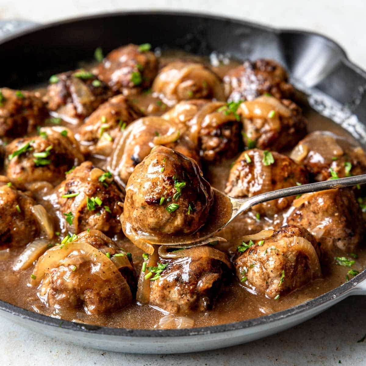 Beef Meatballs in a brown gravy in a skillet with one being ladled out
